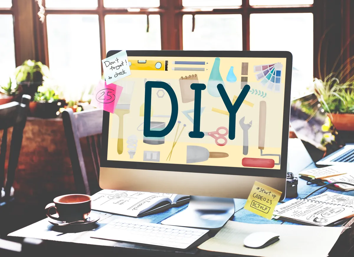 Ordinateur de bureau sur un plan de travail lumineux affichant le mot DIY en grandes lettres bleu marine, entouré d'illustrations d'outils colorés (pinceau, niveau, crayons, clé, ciseaux, nuancier), avec post-it manuscrits, tasse de café et carnets autour : créer son site web soi-même en mode DIY.