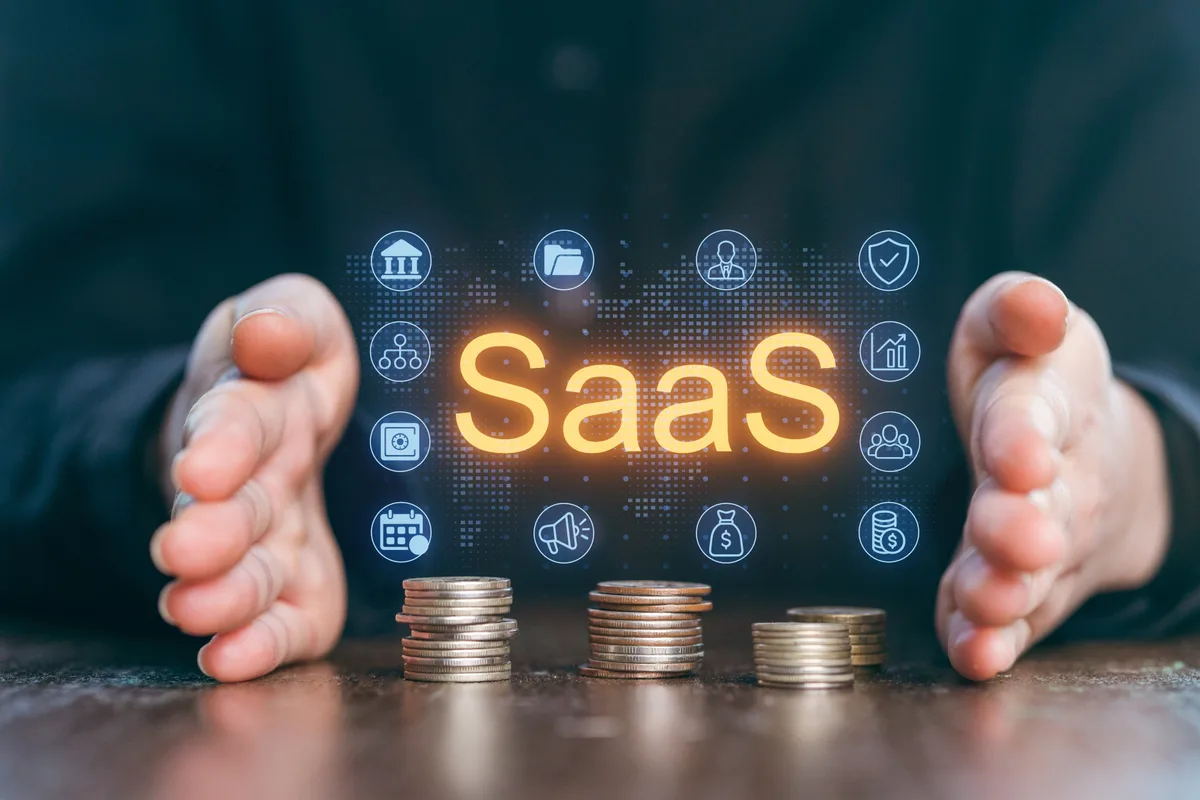 Deux mains encadrent le mot SaaS en lettres lumineuses, entouré d'icônes d'application, au-dessus de trois piles de pièces de monnaie posées sur une table