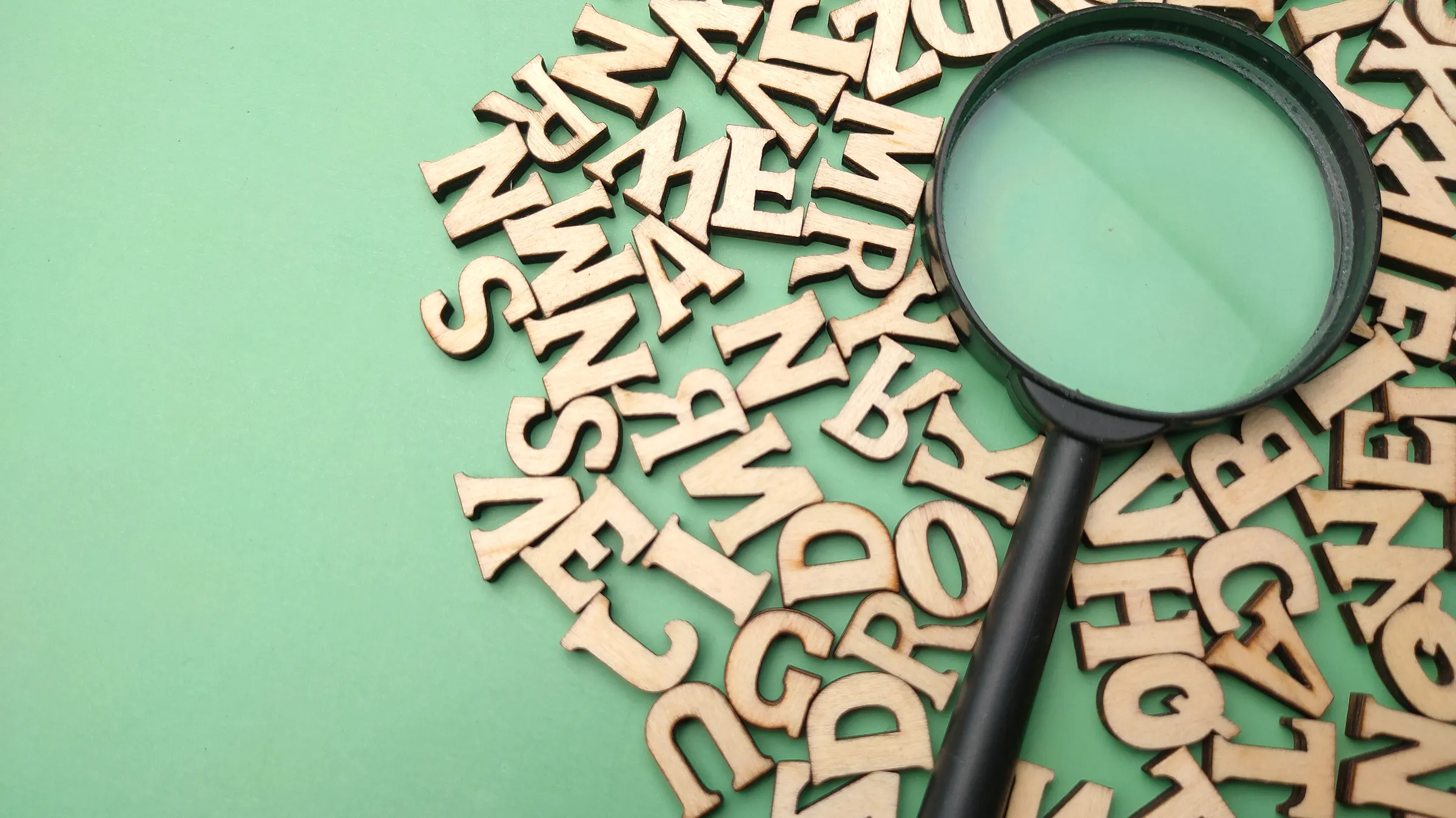 Loupe noire posée sur un tas de lettres en bois éparpillées sur un fond vert, illustrant la recherche des mots-clés que tape vraiment une cible dans Google